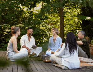 Five adults sit together on a wooden deck outdoors, engaged in conversation at a mental health retreat, surrounded by greenery, with tea and snacks on the ground.