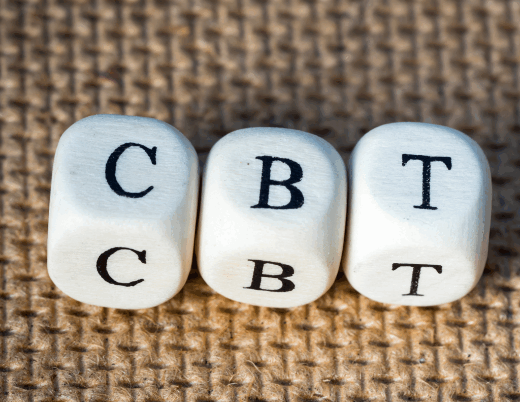 Three white dice with the letters "C," "B," and "T" are lined up on a textured brown surface, representing CBT, also known as Cognitive Behavioral Therapy.