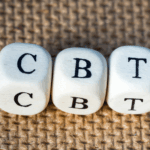 Three white dice with the letters "C," "B," and "T" are lined up on a textured brown surface, representing CBT, also known as Cognitive Behavioral Therapy.