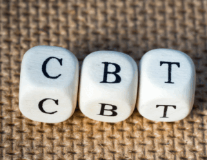 Three white dice with the letters "C," "B," and "T" are lined up on a textured brown surface, representing CBT, also known as Cognitive Behavioral Therapy.