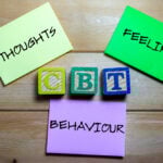 Three note cards labeled "THOUGHTS," "FEELINGS," and "BEHAVIOUR" surround wooden blocks spelling "CBT," representing the core elements of Cognitive Behavioral Therapy on a wooden surface.