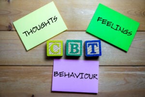 Three note cards labeled "THOUGHTS," "FEELINGS," and "BEHAVIOUR" surround wooden blocks spelling "CBT," representing the core elements of Cognitive Behavioral Therapy on a wooden surface.