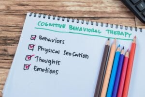 A notebook labeled "Cognitive Behavioral Therapy" (CBT) lists behaviors, physical sensations, thoughts, and emotions, with colored pencils and checked boxes beside each item.
