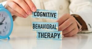 A person arranges three wooden blocks labeled "Cognitive," "Behavioral," and "Therapy" on a white surface next to a blue alarm clock, symbolizing Cognitive Behavioral Therapy (CBT) and its focus on timely interventions.