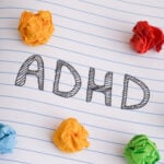 The word "ADHD" is written on a lined notebook page, surrounded by crumpled balls of colorful paper, highlighting the daily challenges faced by those with Attention Deficit Disorder.
