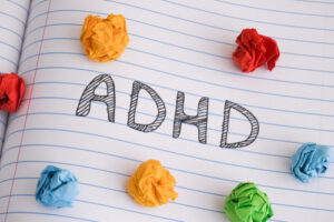 The word "ADHD" is written on a lined notebook page, surrounded by crumpled balls of colorful paper, highlighting the daily challenges faced by those with Attention Deficit Disorder.