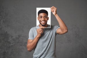 A man in a gray t-shirt holds a smiling photo of his face in front of his head, standing against a gray textured background, representing how some unmask ADHD or hide ADHD symptoms behind a cheerful facade.