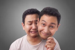 A man with a sad facial expression holds a mask of his own happy face in front of him, symbolizing ADHD masking, against a gray background.
