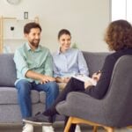 A couple sits together on a couch, smiling and holding hands, while talking to a professional during relationship counseling in Los Angeles.