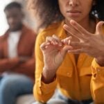 A woman sits on a couch removing a ring from her finger, while a man in the background sits with his arms crossed—a powerful moment reflecting the stages of a dying marriage.
