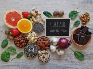 A variety of foods including citrus fruits, nuts, seeds, spinach, strawberry, garlic, onion, and dark chocolate surrounds a sign reading "CORTISOL Lowering foods," showcasing evidence-based natural remedies to help treat anxiety.