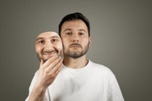 A man in a white shirt holds a smiling mask in front of his face, revealing a neutral expression underneath—a striking depiction of ADHD masking and the effort to hide true feelings.