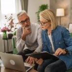 An older man and woman sit on a green couch, looking at documents and a laptop, appearing to discuss finances in relationships in their well-lit living room.