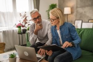 An older man and woman sit on a green couch, looking at documents and a laptop, appearing to discuss finances in relationships in their well-lit living room.